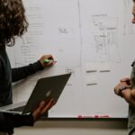 Two people discussing a performance analytics strategy on a whiteboard while one holds a laptop