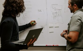Two people discussing a performance analytics strategy on a whiteboard while one holds a laptop