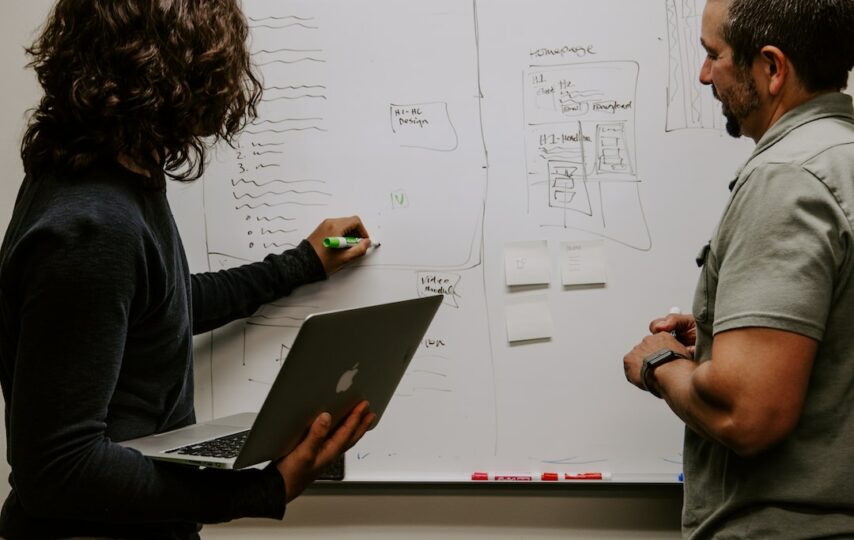 Two people discussing a performance analytics strategy on a whiteboard while one holds a laptop